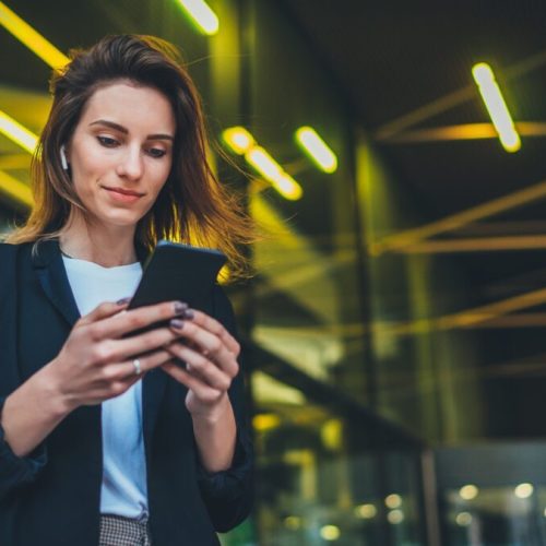 Successful female banker using smartphone outdoors while standing near his office background yellow neon lights, young woman professional manager working on mobile device near skyscraper at night